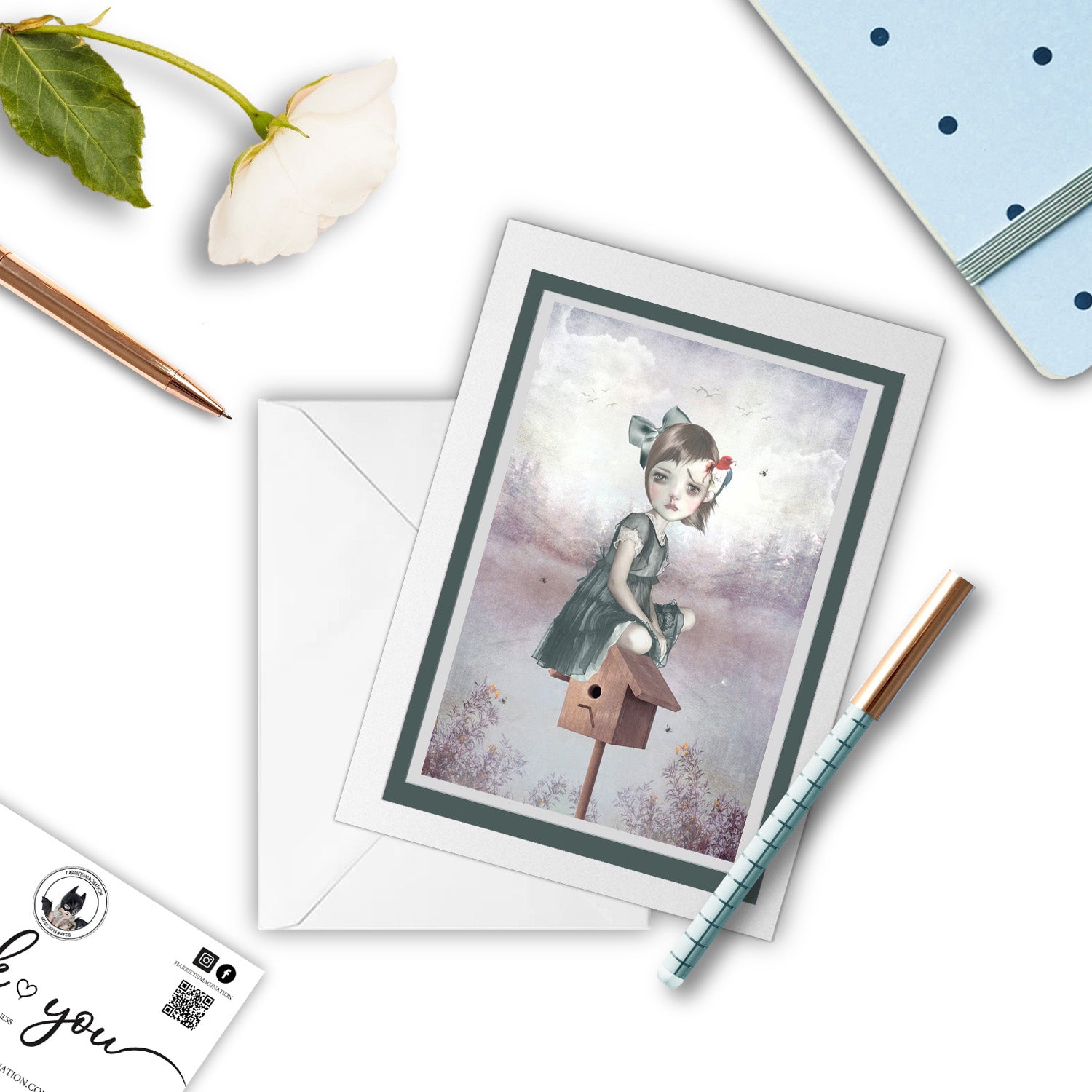 Art greeting card featuring a girl and woodpecker, displayed alongside a book and pen.