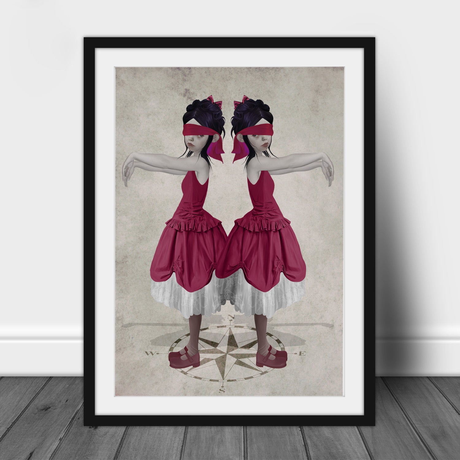 Framed art print of twin sisters blindfolded on a compass, displayed in a black frame leaning against a  white background.