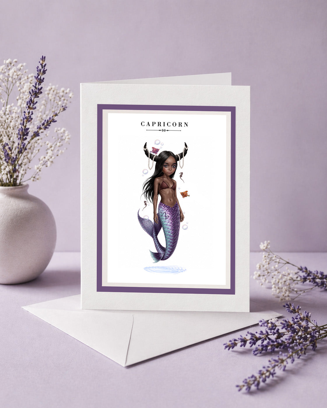 Capricorn zodiac birthday card featuring an illustration of a mermaid with dark skin, goat horns draped in pearls, and a iridescent purple fish tail.