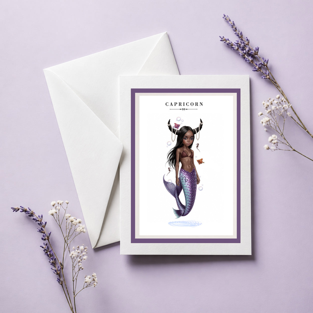 Capricorn zodiac birthday card and envelope featuring an illustration of a mermaid with dark skin, goat horns draped in pearls, and a iridescent purple fish tail.