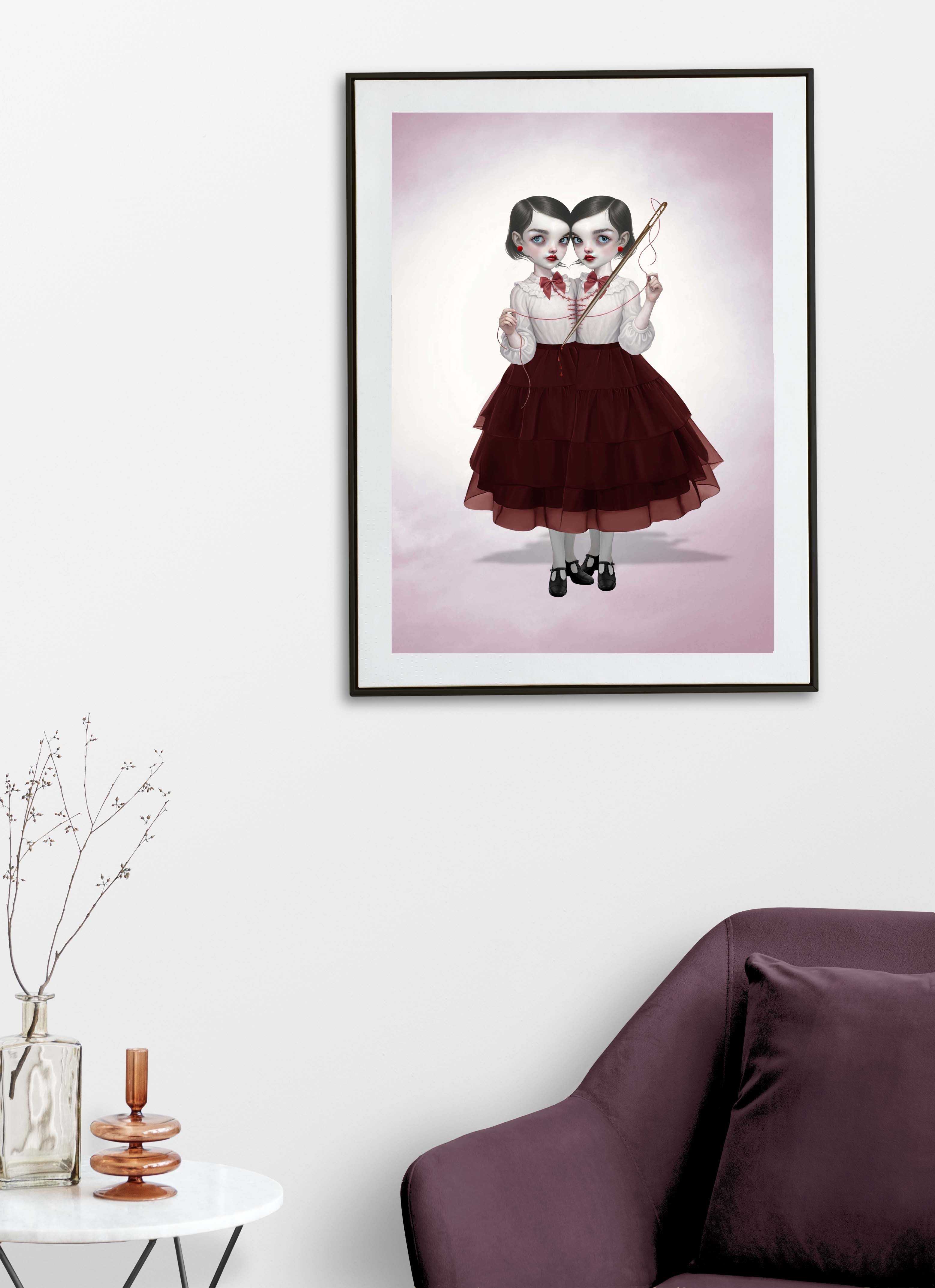 Dark pop surrealism art print on a living room wall above chair. This gothic illustration features conjoined twin girls with a needle and thread.