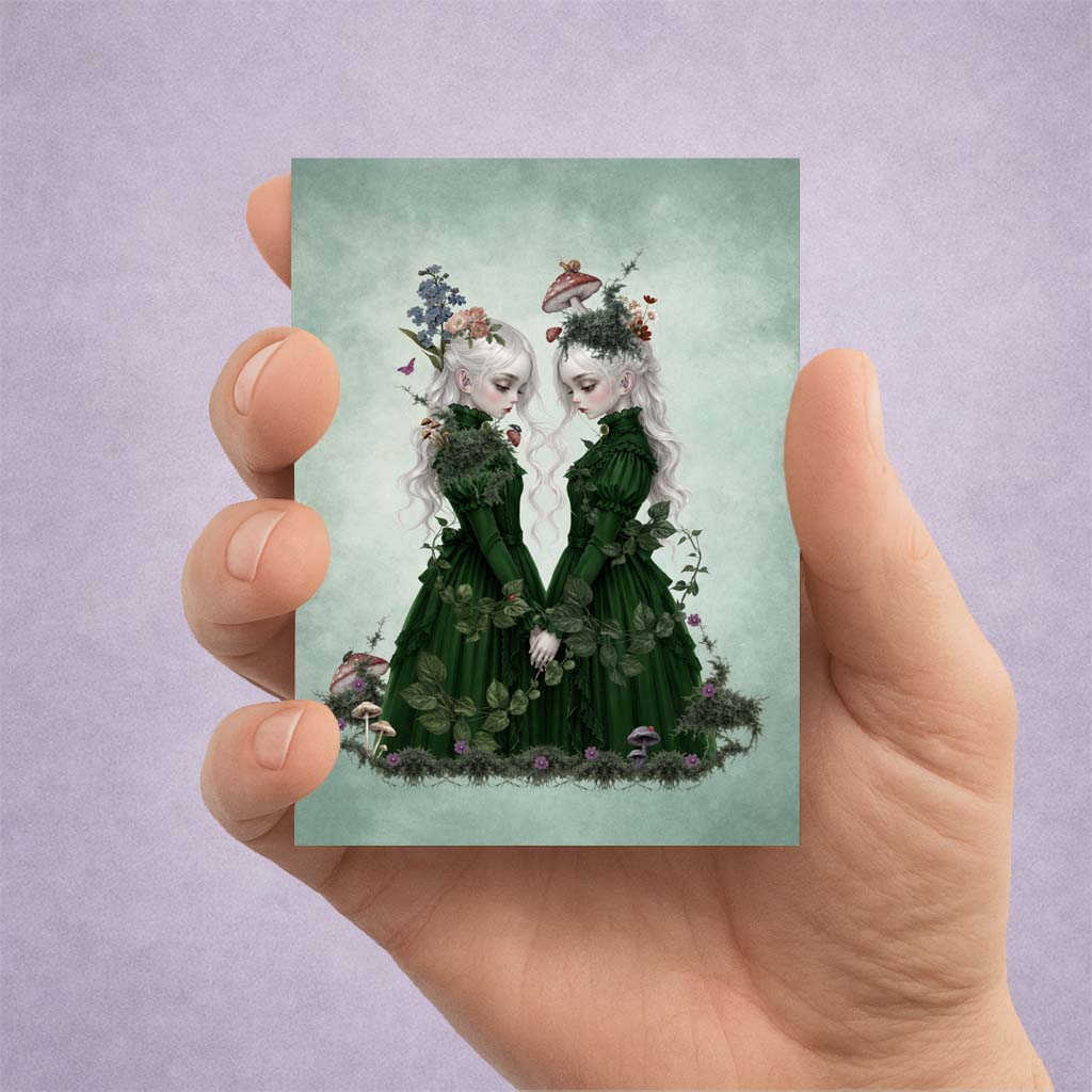 A hand holding a miniature 2.5 by 3.5 inch ACEO art card print featuring two ethereal twin girls with white hair in dark green Victorian dresses.