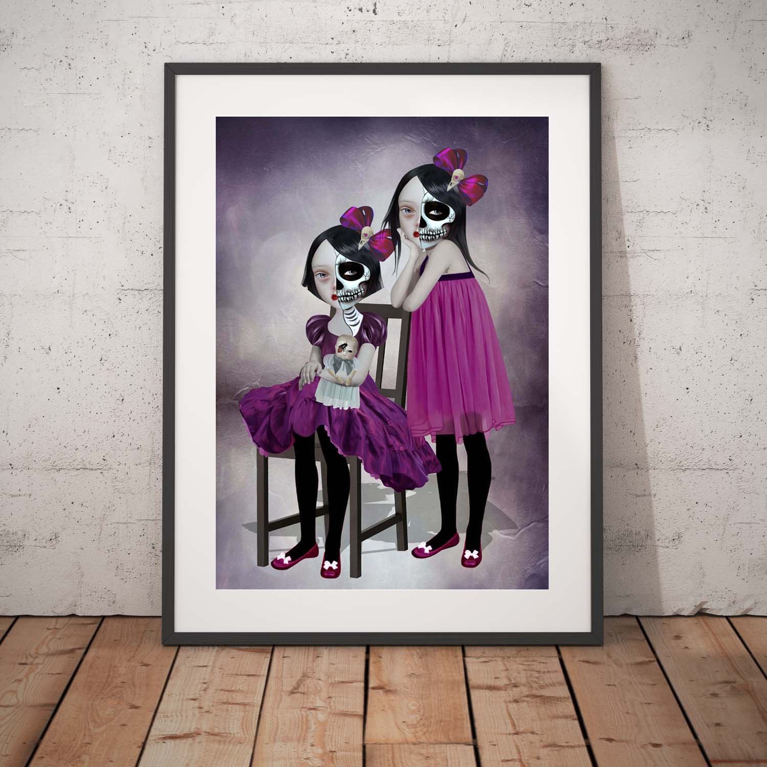 Day of the dead sisters art print in a black frame, standing against a white wall.