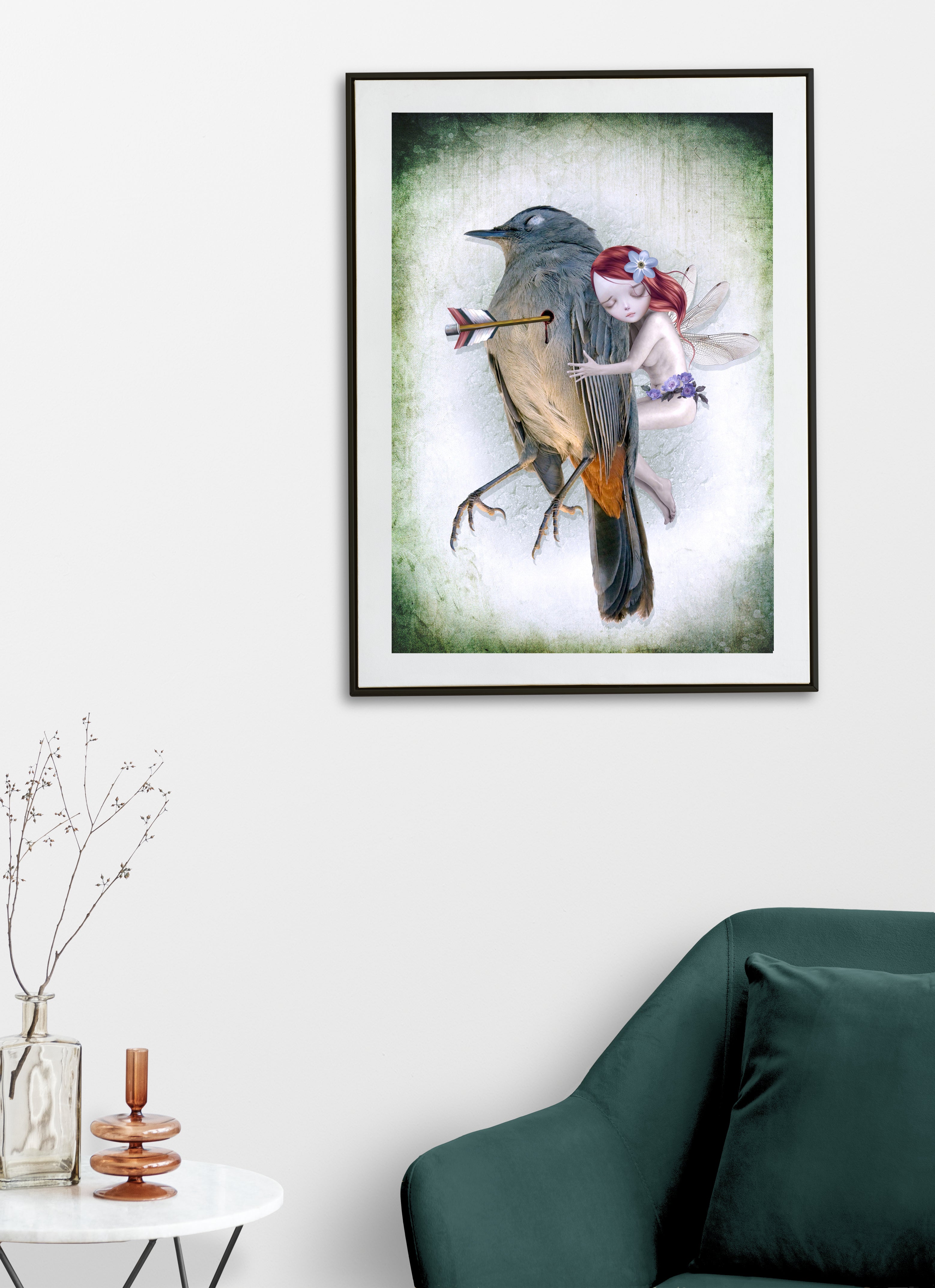 Framed artwork of a fairy hugging a wounded song bird on a white wall above a green sofa.
