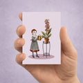 Hand holding an ACEO card with an illustration of a girl watering Foxglove flowers with real fox heads against a light purple background