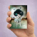 Hand holding a card with an artistic illustration of a woman with dark hair and butterflies, against a purple background.