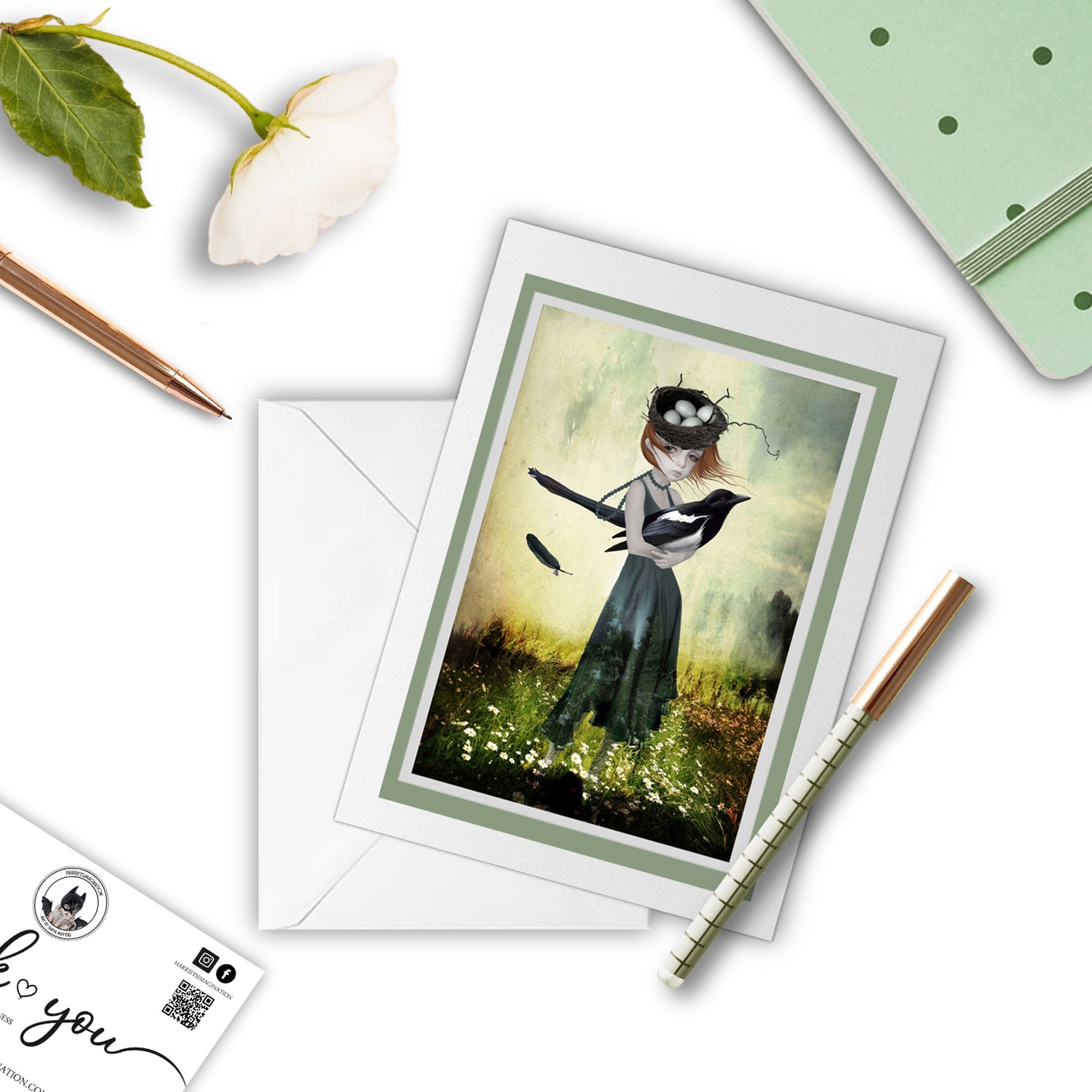 Girl and magpie art greeting card, with a pen and other stationaryitems arranged around it.