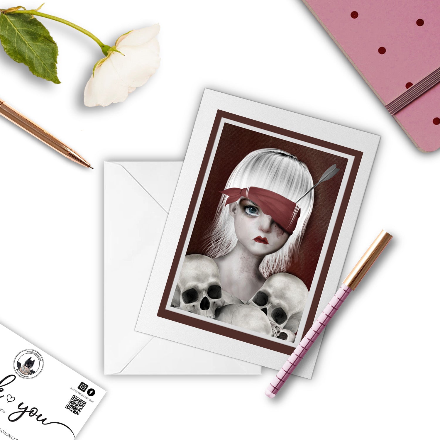 Macabre Girl and Skulls Art Greeting Card with a pen and other stationary  items arranged around it.