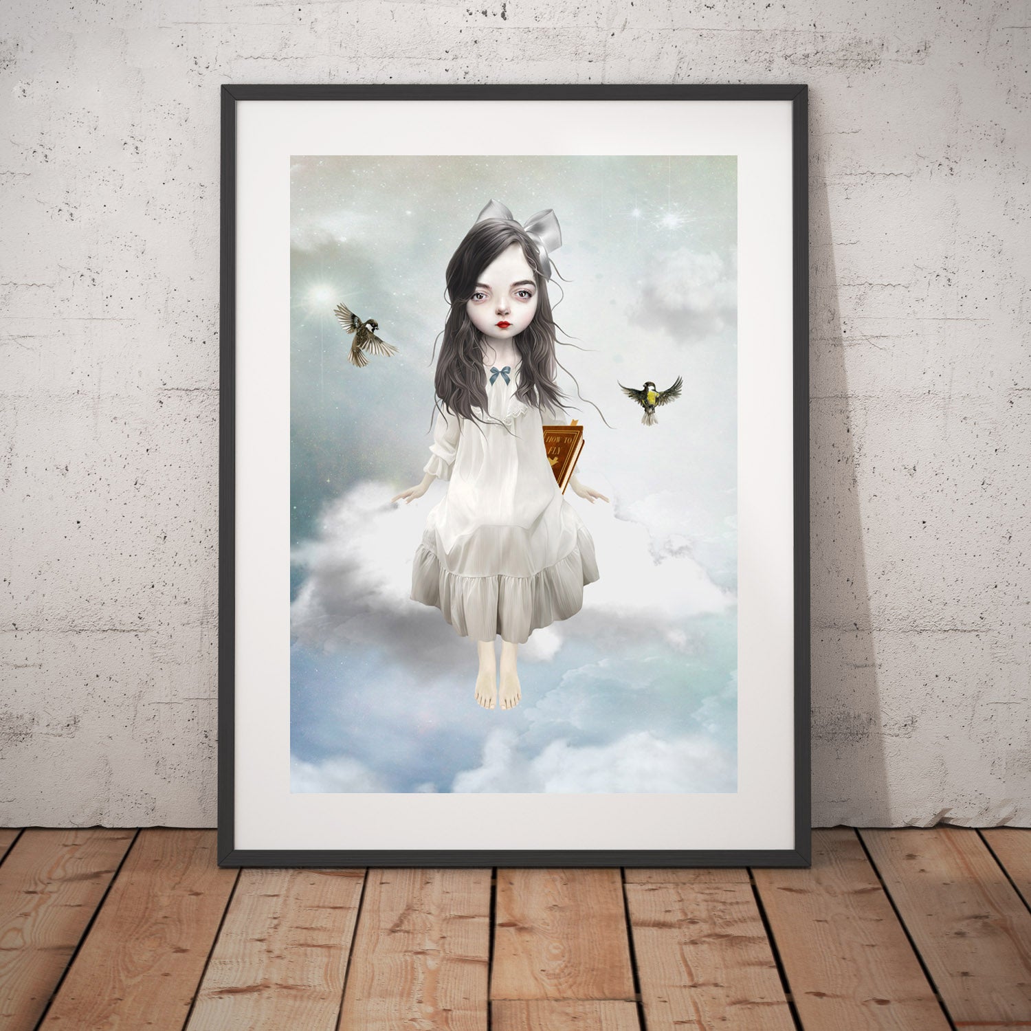 Girl sitting on a cloud art print in a black frame, standing against a white wall.