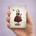 Hand holding a card with an illustration of a girl in a forest surrounded by beetles and butterflies.
