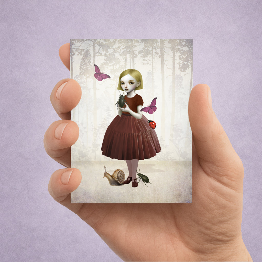 Hand holding a card with an illustration of a girl in a forest surrounded by beetles and butterflies.
