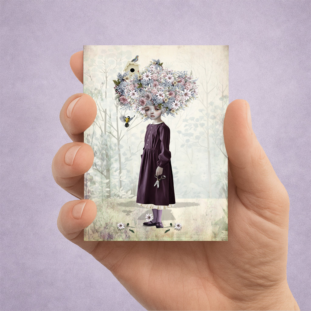 Hand holding a card with a design of a girl with flowers as her hair against a light background.