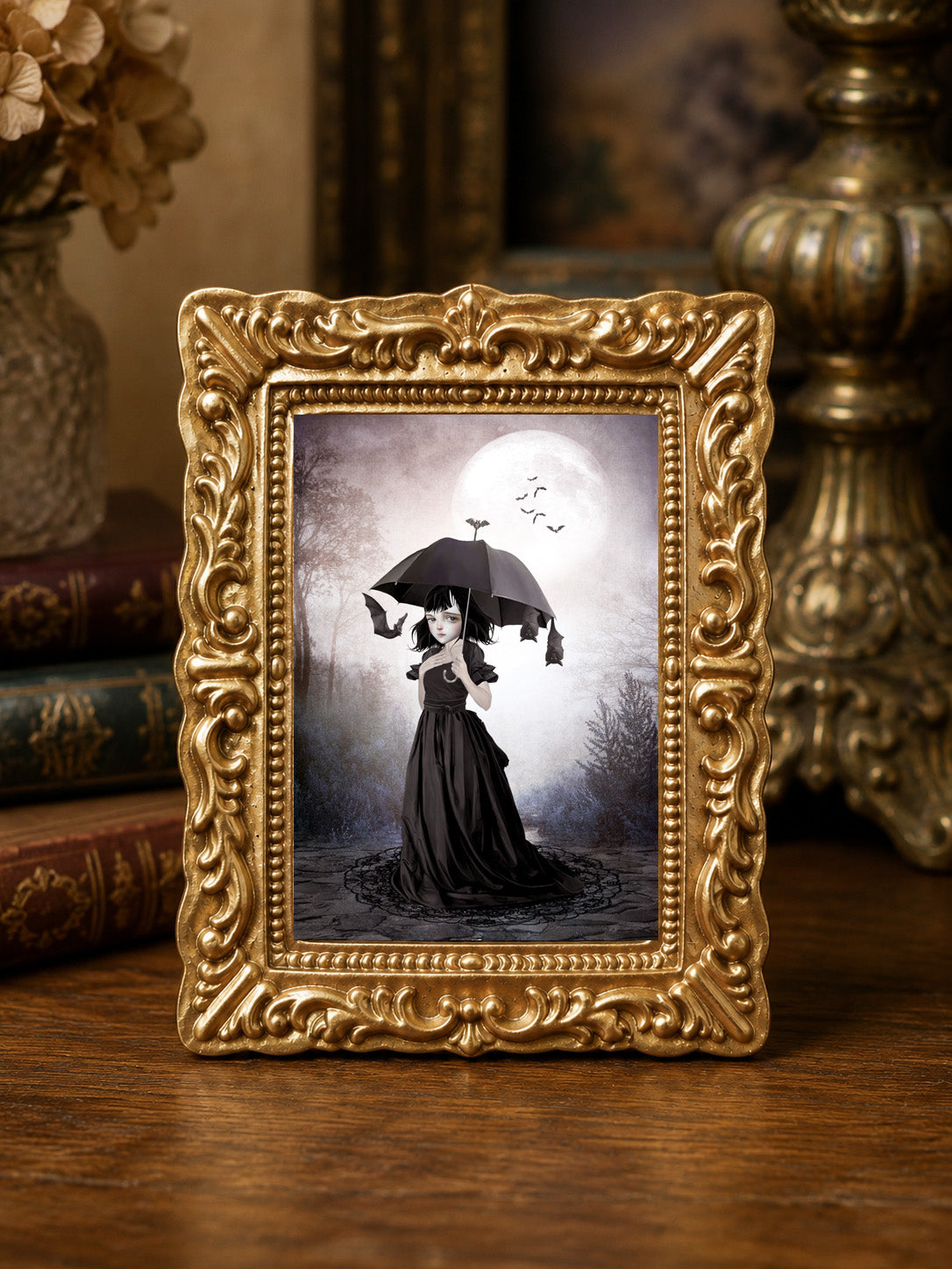 Miniature 1:12 scale gothic art print in an ornate gold frame featuring a young girl in a black Victorian dress holding an umbrella, surrounded by hanging bats beneath a full moon, displayed in a dollhouse setting.