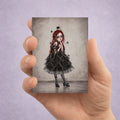 ACEO Card held in hand showing a melancholy girl with vibrant red hair wearing a dark ruffled dress and sheer tights, with intricate cobwebs over her face and black spiders hanging around her