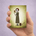 Hand holding an ACEO card with an illustration of a girl holding a flower against a purple background.