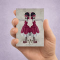 Hand holding an ACEO card with an illustration of twin girls in red dresses standing on a compass 
