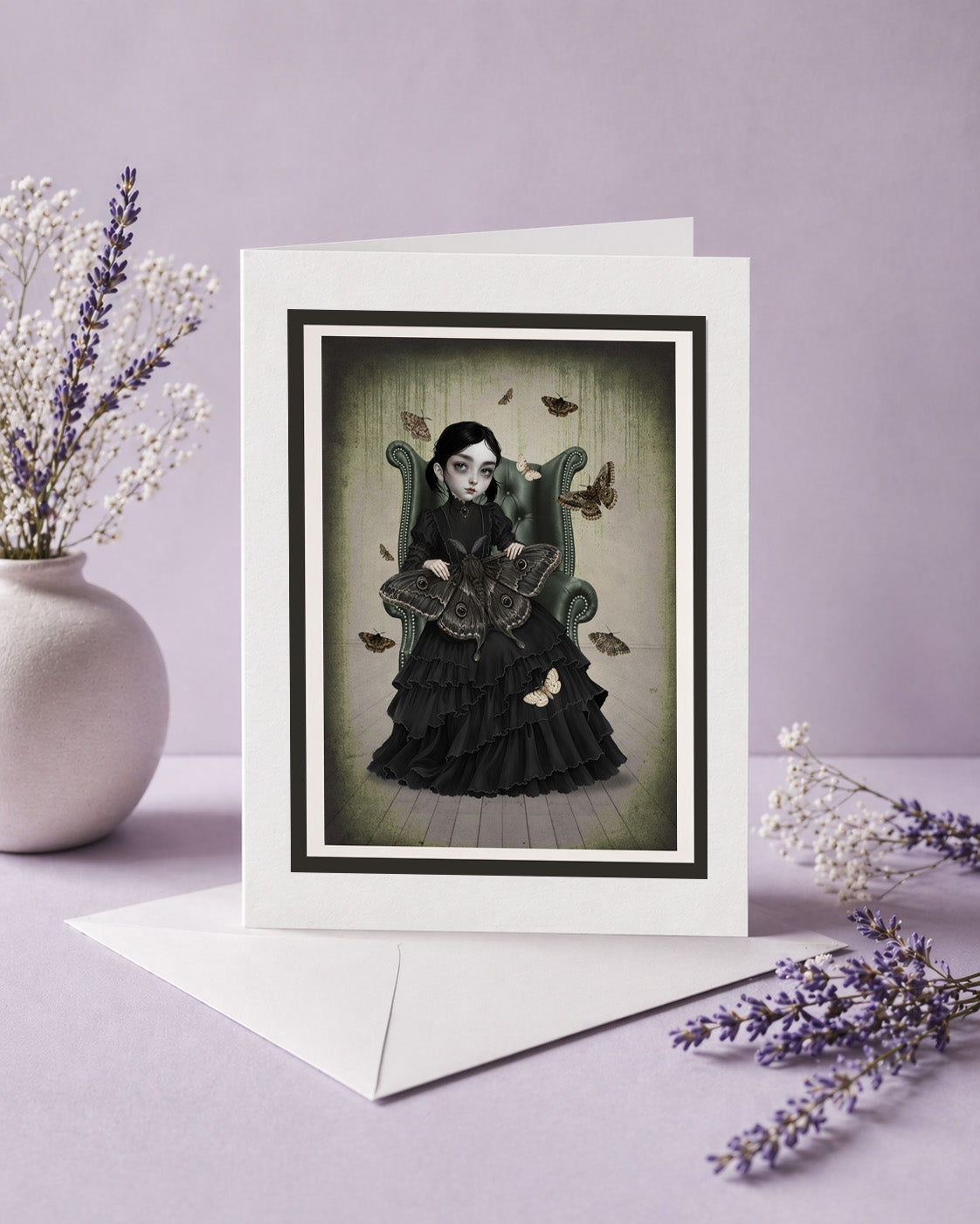 Vertical blank art greeting card featuring a gothic girl with moths sitting in an armchair, displayed upright next to a white envelope and dried lavender flowers.