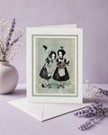 Blank greeting card with white envelope displayed on a purple background, featuring twin gothic girls with blackbirds and a pie in a dark cottagecore style.