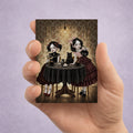 2.5x3.5 inch ACEO miniature art card held in hand featuring two Victorian gothic girls having a dark tea party with a black cat, voodoo doll, and playing cards.