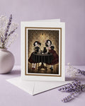 A greeting card with a dark gothic tea party artwork framed in gold, displayed on a purple backdrop with dried lavender and a white envelope