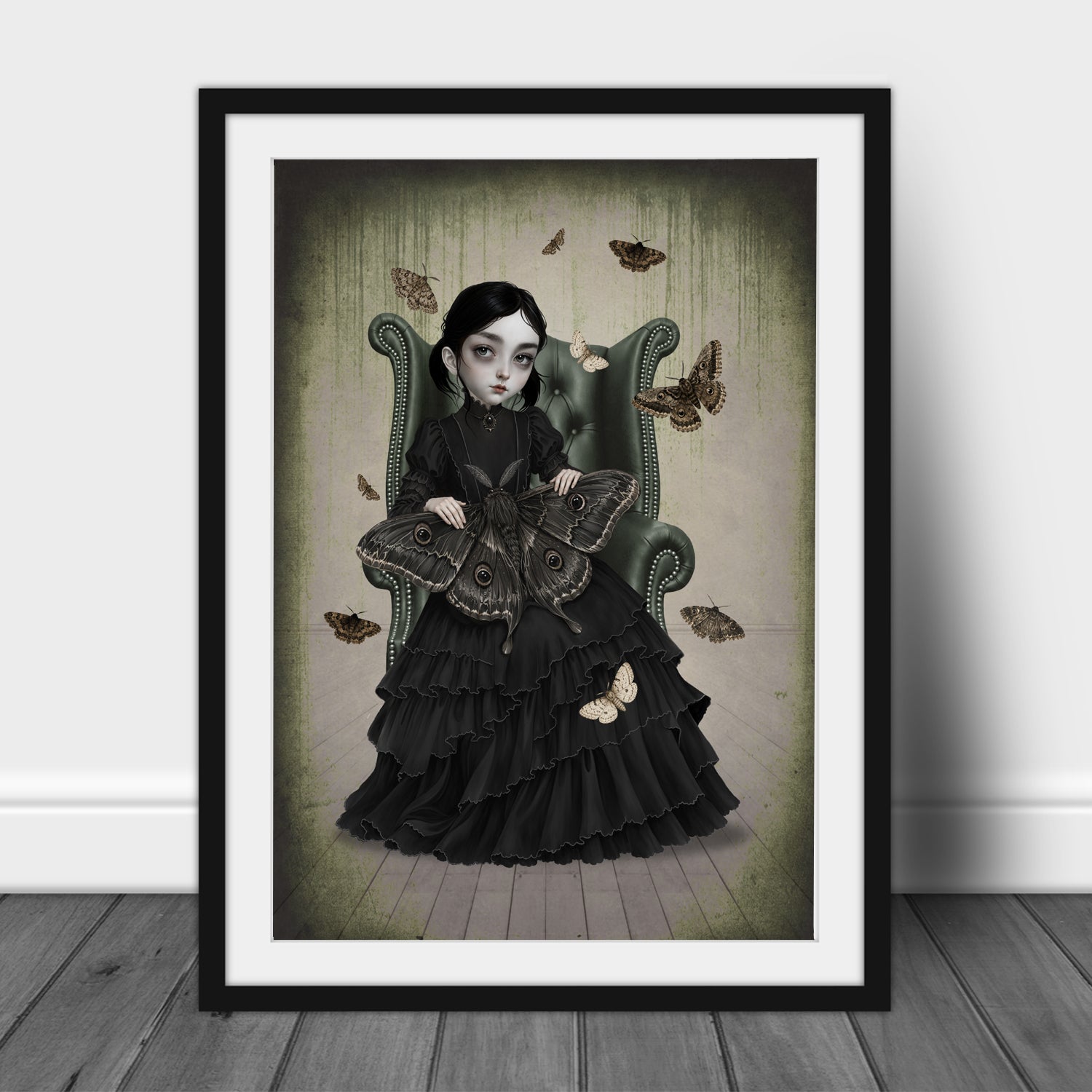 Gothic girl in a black Victorian ruffled dress sitting on a green armchair holding a giant moth, surrounded by fluttering moths in a dark cottagecore art print displayed in an A3 frame