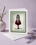 A white greeting card with a dark green border and an illustration of a girl in a red dress holding a anatomical heart, displayed with a white envelope on a lavender surface with dried flowers.