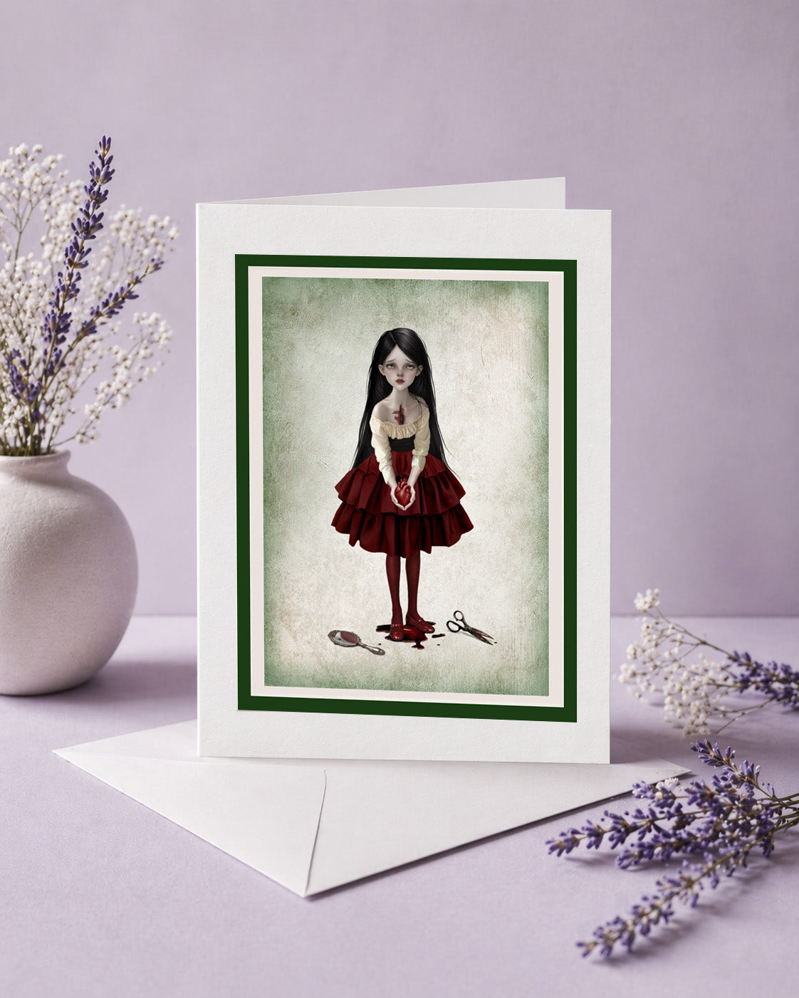 A white greeting card with a dark green border and an illustration of a girl in a red dress holding a anatomical heart, displayed with a white envelope on a lavender surface with dried flowers.