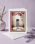 Greeting card with a vintage-style illustration of a headless clown in a circus setting, surrounded by lavender flowers on a light purple background.