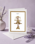 Libra zodiac birthday card featuring an illustration of a blonde maiden in a metallic gold dress sitting atop a golden balance scale with a tiny bird.
