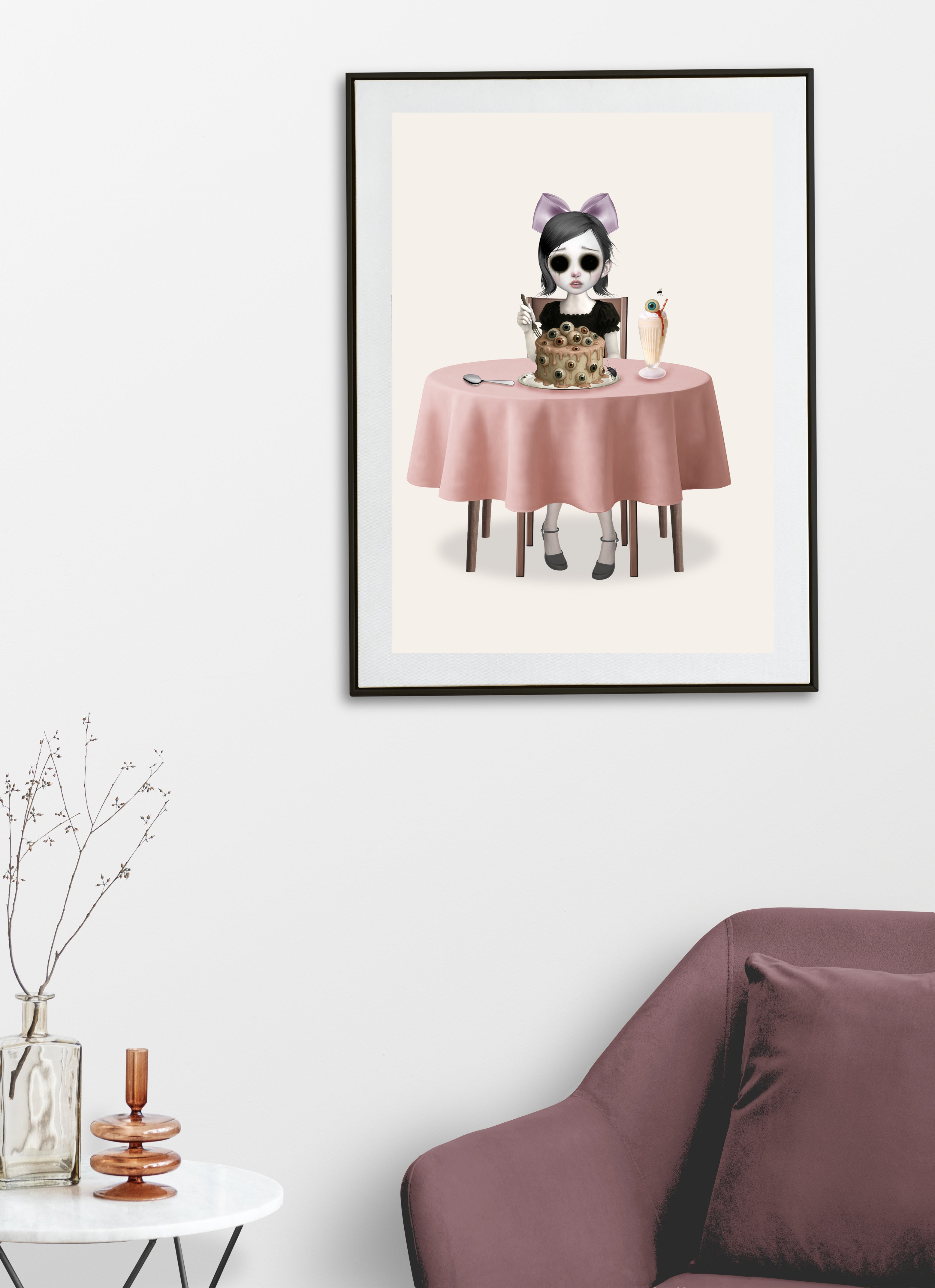 Surreal pastel goth wall decor featuring a framed print of an eyeball cake illustration styled above a chair on a clean white wall.