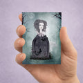 Hand holding an ACEO card with a gothic design of a ghost girl by her tombstone against a purple background.