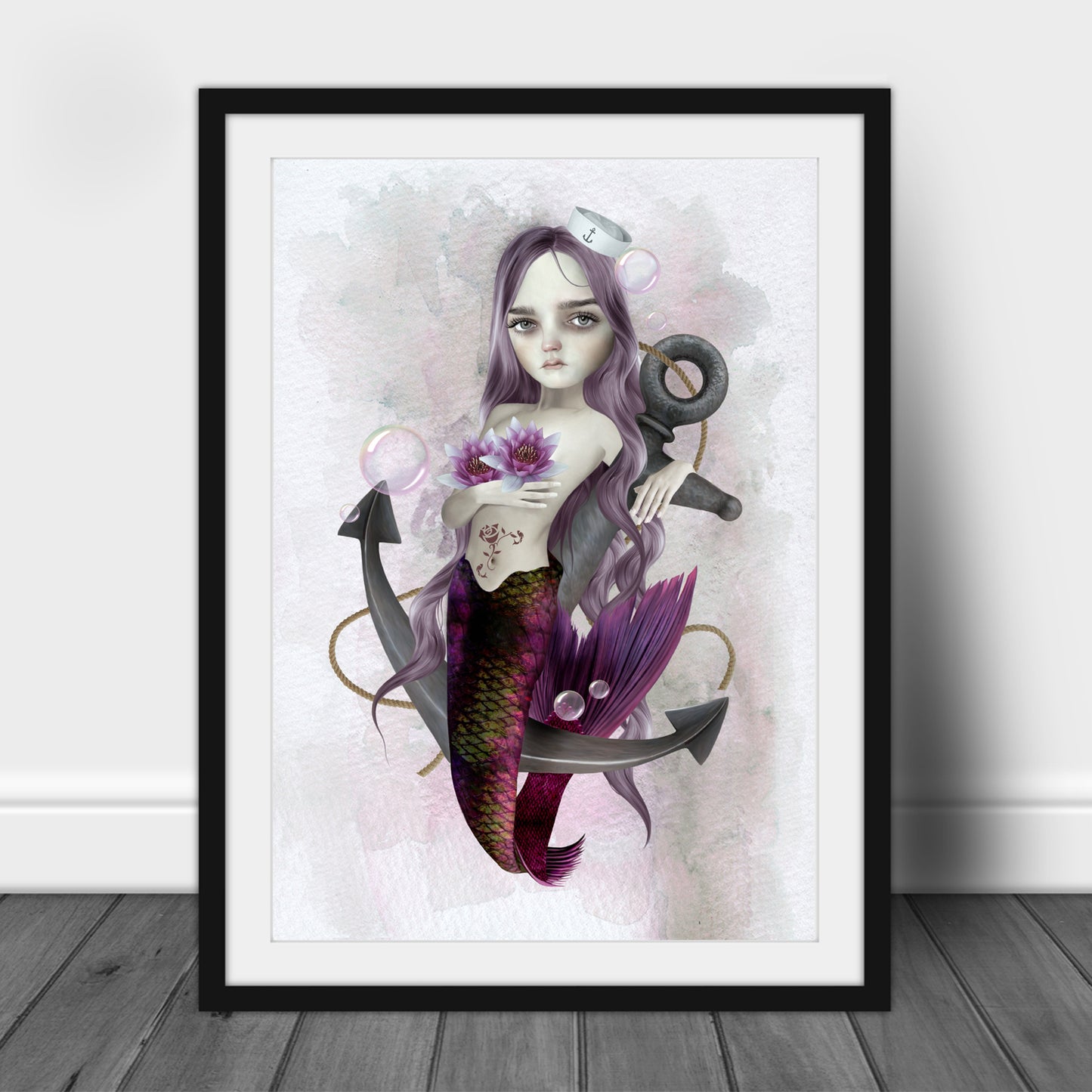 Mermaid and anchor print in a black frame, standing against a white wall.
