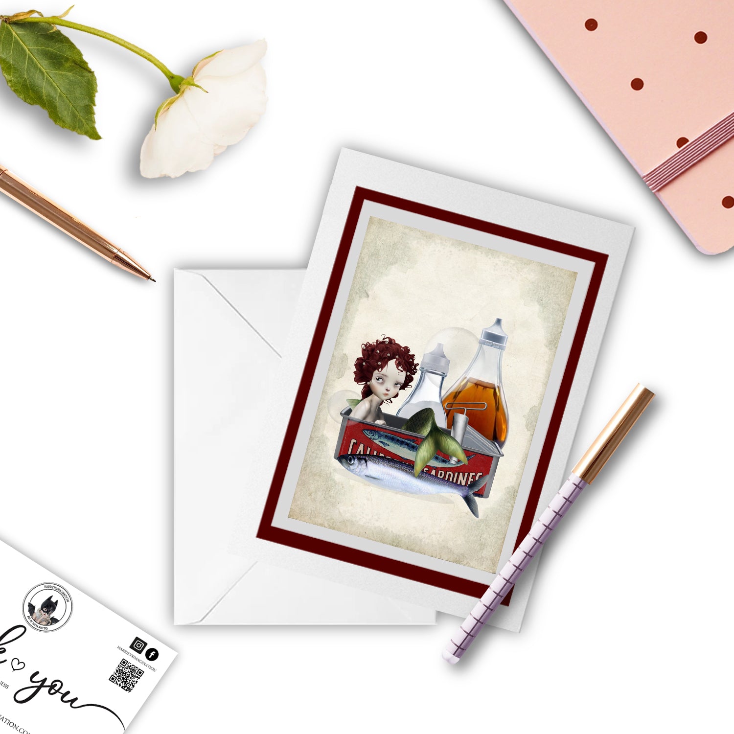 Mermaid in sardine tin art greeting card, with a pen and other stationary items arranged around it.