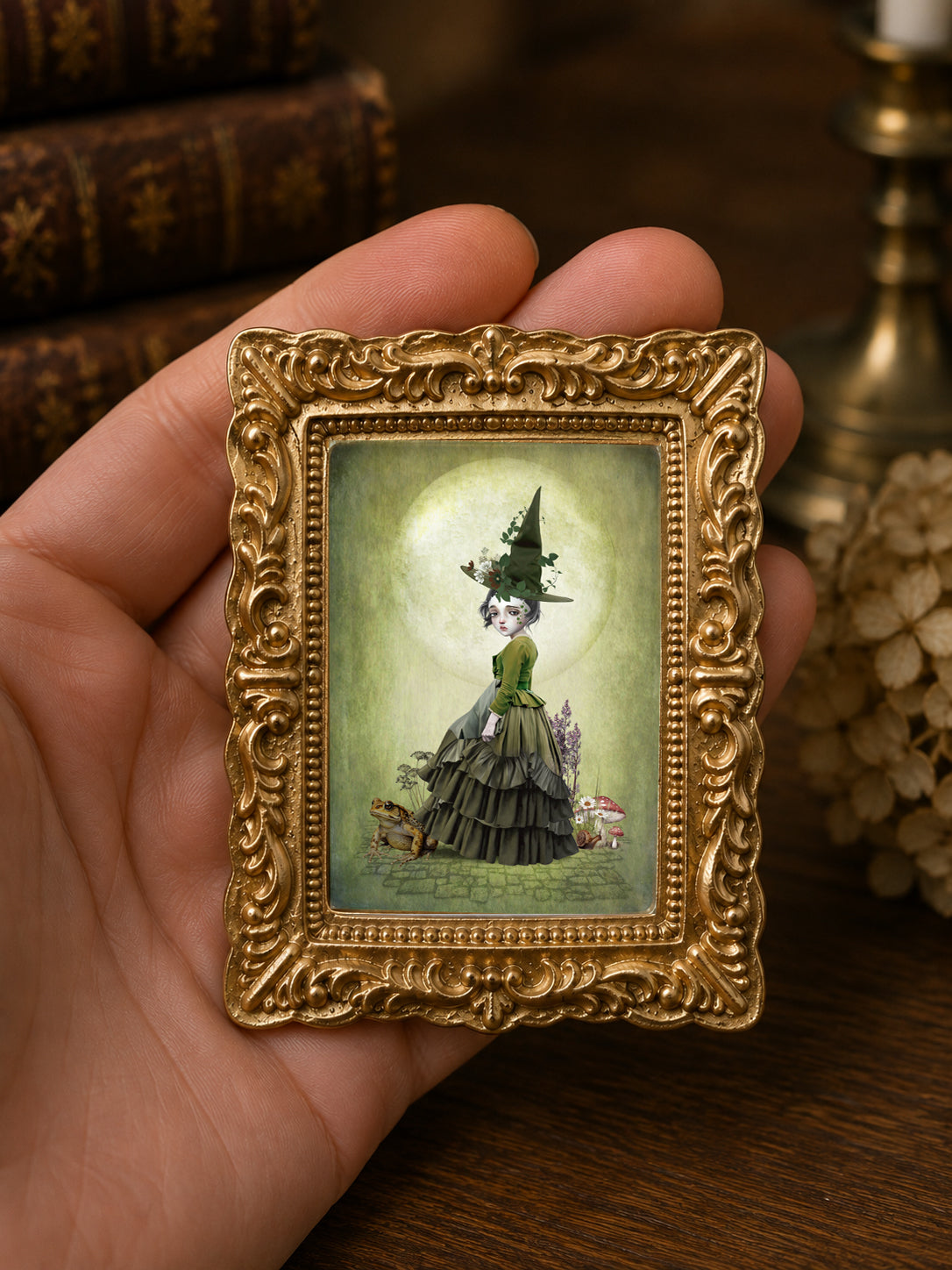 Miniature 1:12 scale cottagecore witch art print in an ornate gold frame featuring a young witch in a green dress and pointed hat, standing with a large toad and mushrooms under a full moon, displayed in a dollhouse setting.