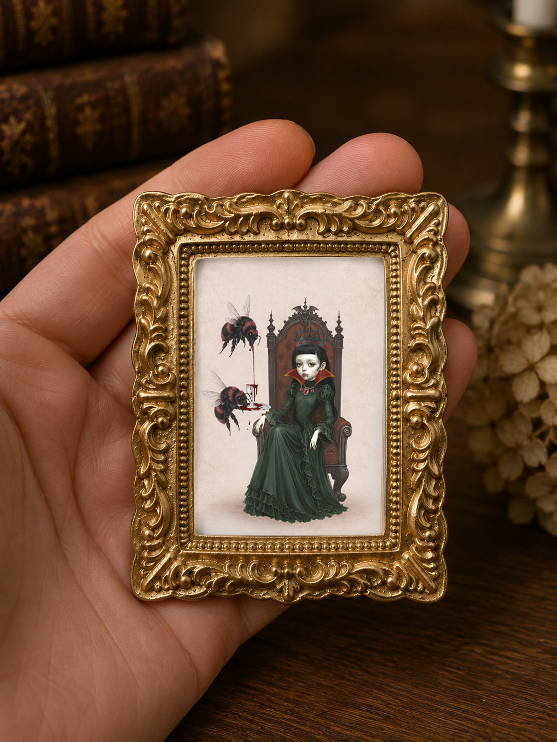 Miniature 1:12 scale gothic art print in an ornate gold frame featuring a dark queen on a throne with red bumblebees pouring blood into a cup, displayed in a dollhouse setting.