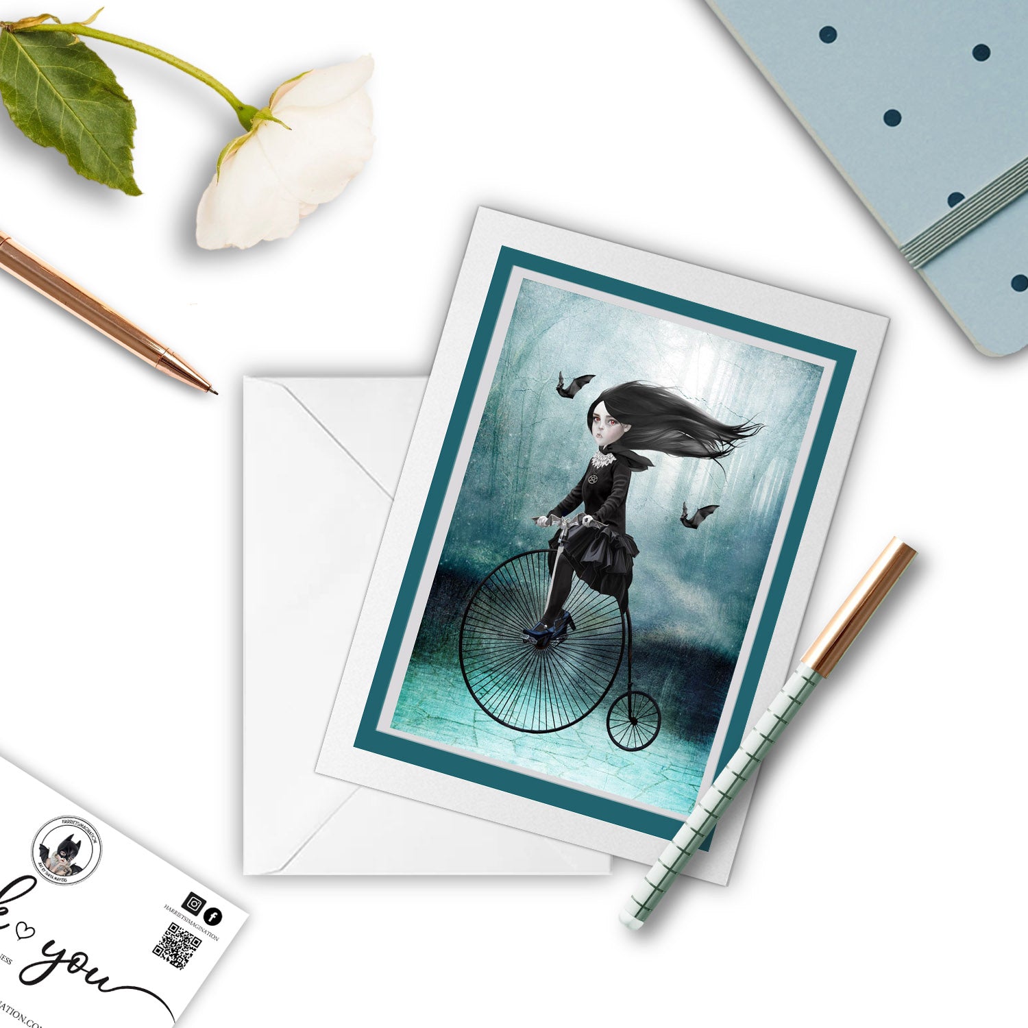 Mockup of vampire on penny farthing greeting card, with a pen and other stationary items arranged around it.
