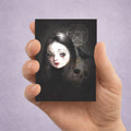 A hand holding an ACEO art card featuring a dark gothic illustration of a pale-faced girl with long dark hair, a skull, a blackbird, and a pentagram on a black background, set against a soft lavender studio backdrop.