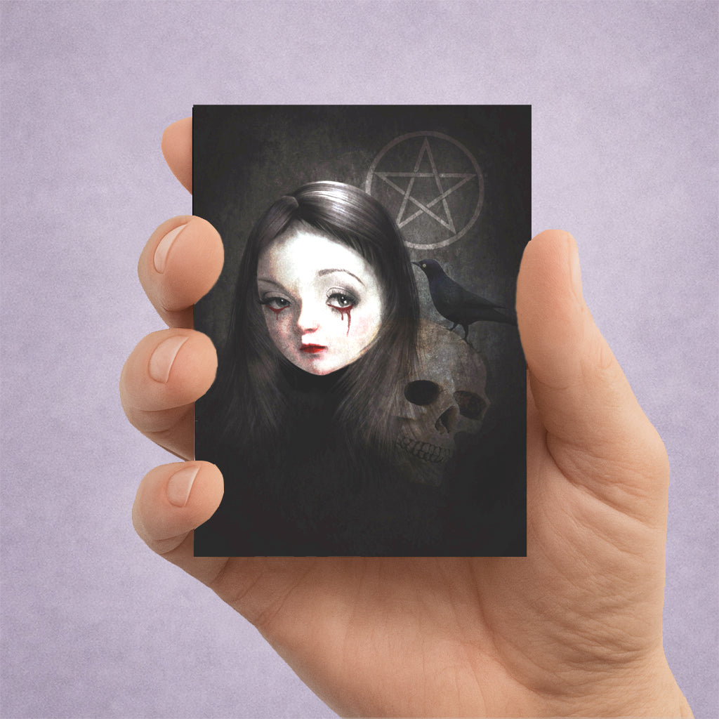 A hand holding an ACEO art card featuring a dark gothic illustration of a pale-faced girl with long dark hair, a skull, a blackbird, and a pentagram on a black background, set against a soft lavender studio backdrop.