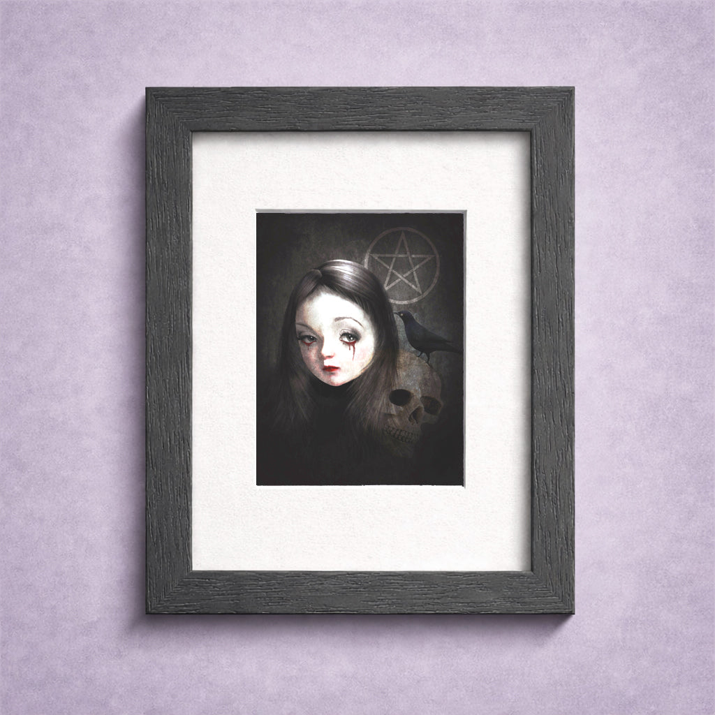 A framed ACEO art Card featuring a gothic illustration of a girl with dark hair, a blckbird, and a skull with a pentagram symbol, set in a dark gray frame with a white mat against a textured light purple wall.