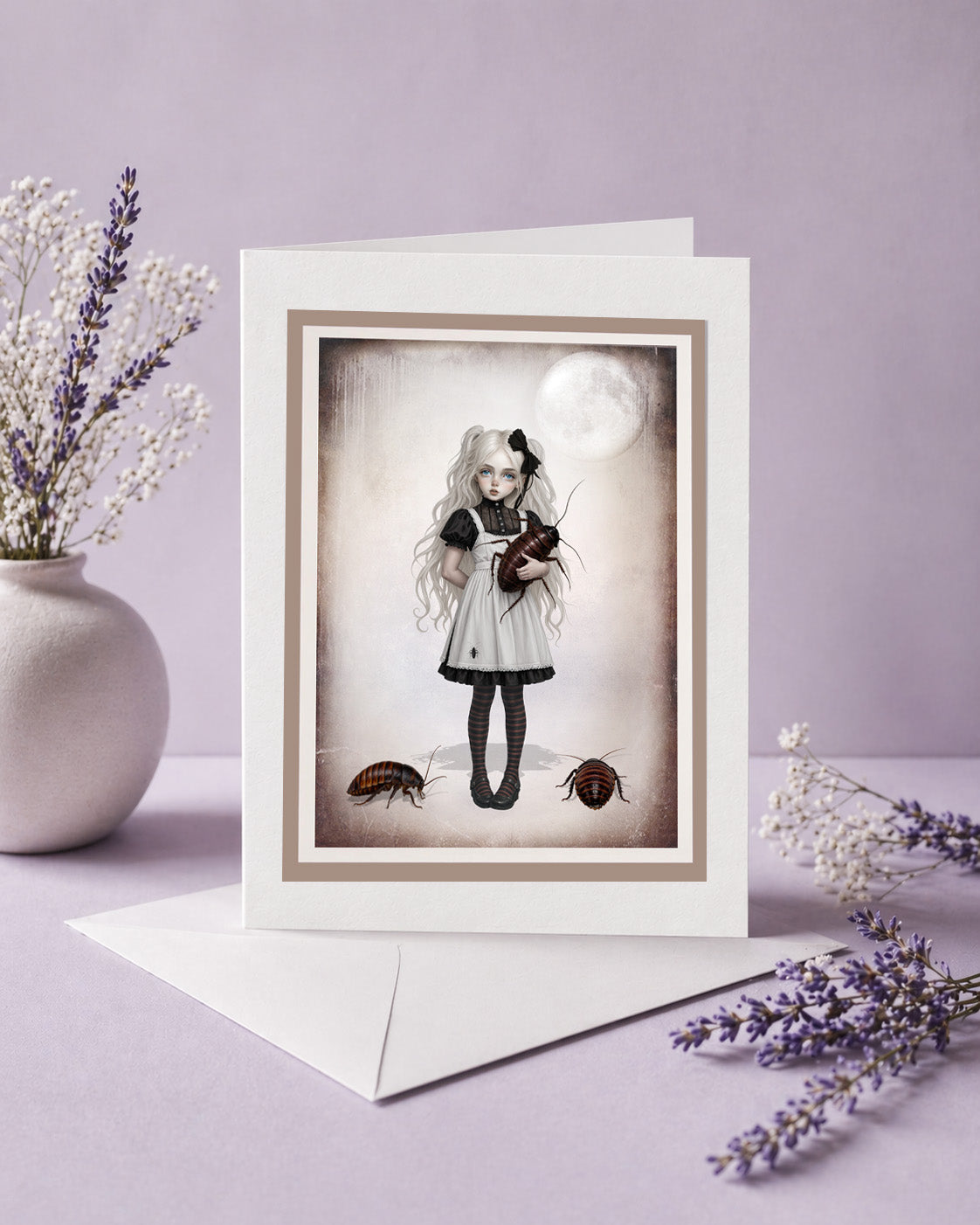 A standing greeting card resting on an envelope, featuring dark surrealism art of a gothic girl holding a giant cockroach, surrounded by lavender sprigs