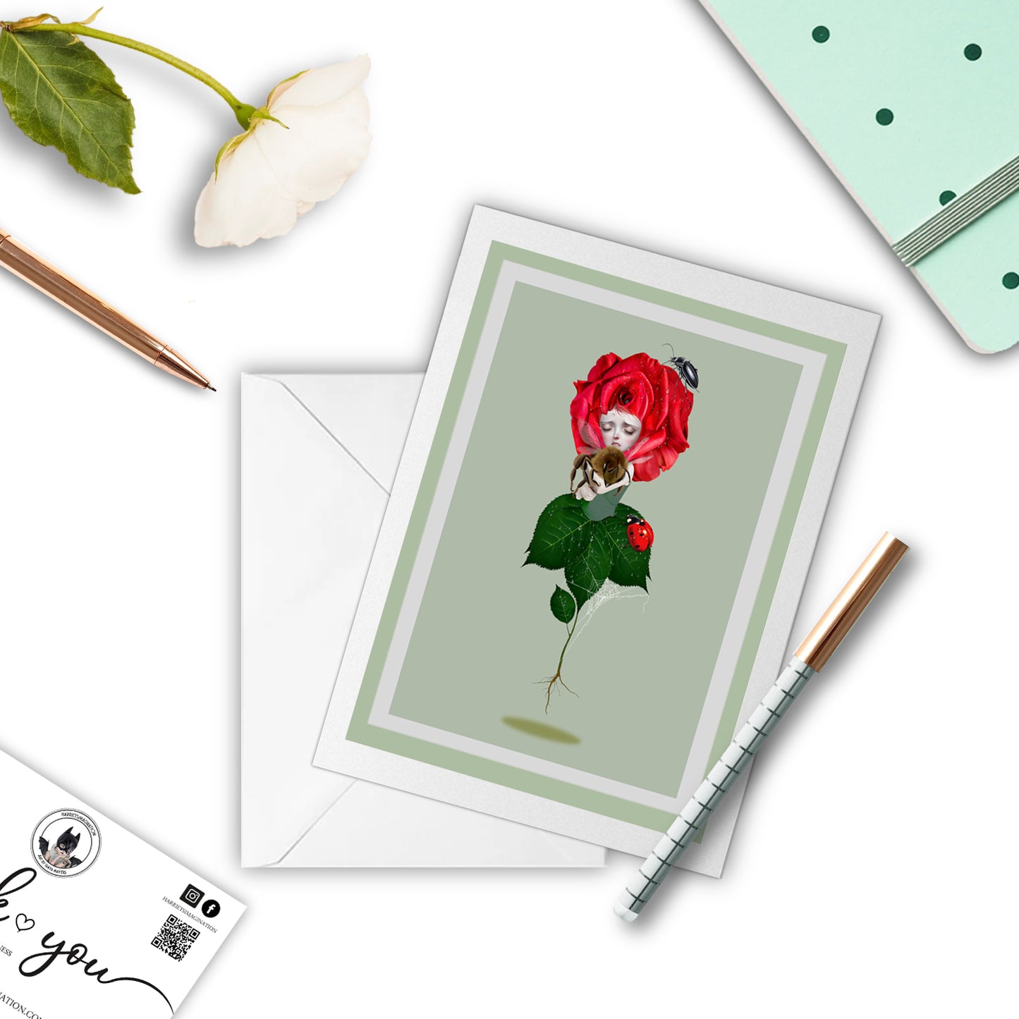 Red Rose And Bumblebee Art Greeting Card with a pen and other stationary items arranged around it.