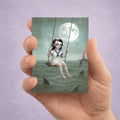 Hand holding a card with an illustration of a sailor girl on a swing over shark infested waters under a moonlit sky.