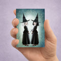 Miniature 2.5x3.5 ACEO art card held in hand, featuring twin gothic witches with white and black hair against a celestial teal background.