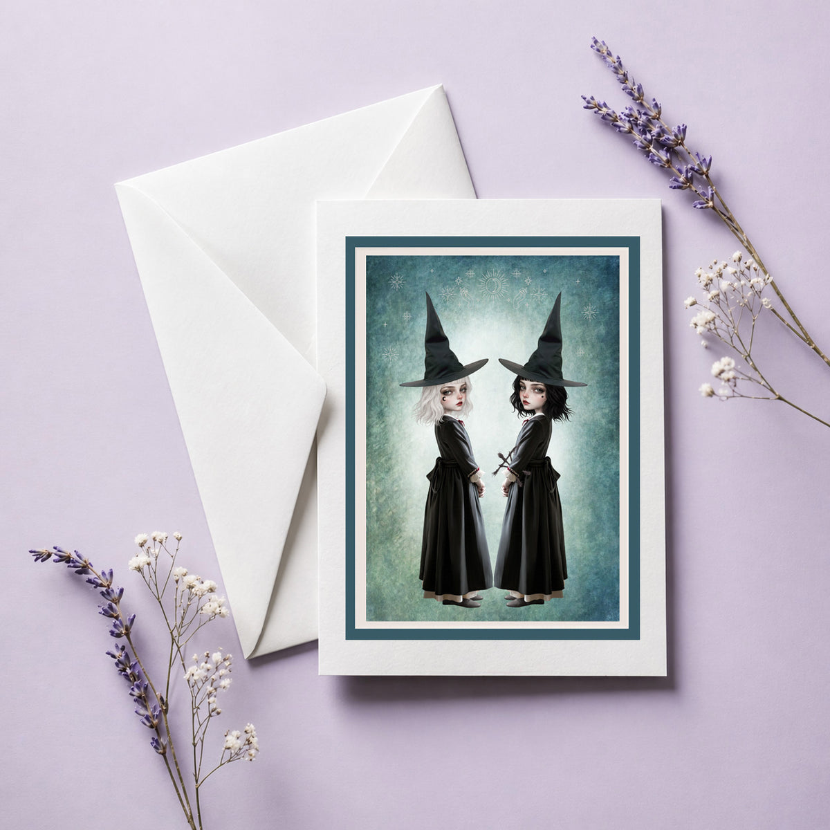Blank gothic art greeting card featuring twin witch sisters in black pointed hats standing back to back against a celestial teal background, displayed with a white envelope.