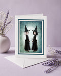 Creepy cute art greeting card with envelope featuring twin witch sisters in black pointed hats standing back to back against a celestial teal background.