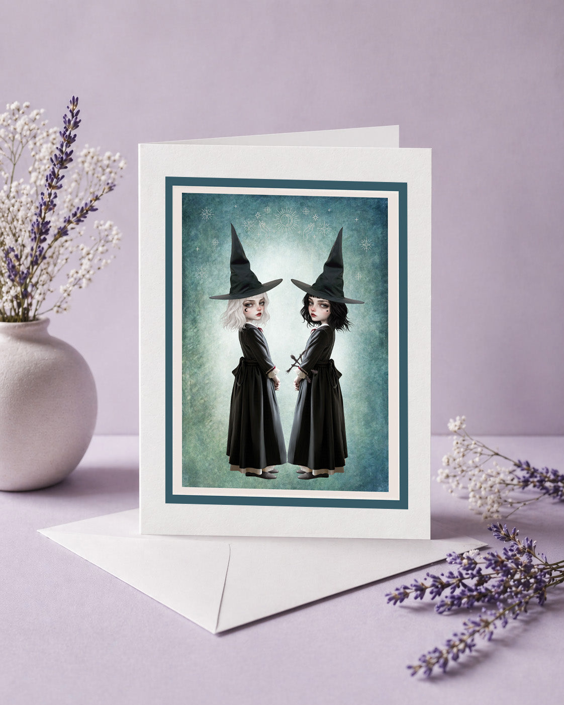 Creepy cute art greeting card with envelope featuring twin witch sisters in black pointed hats standing back to back against a celestial teal background.
