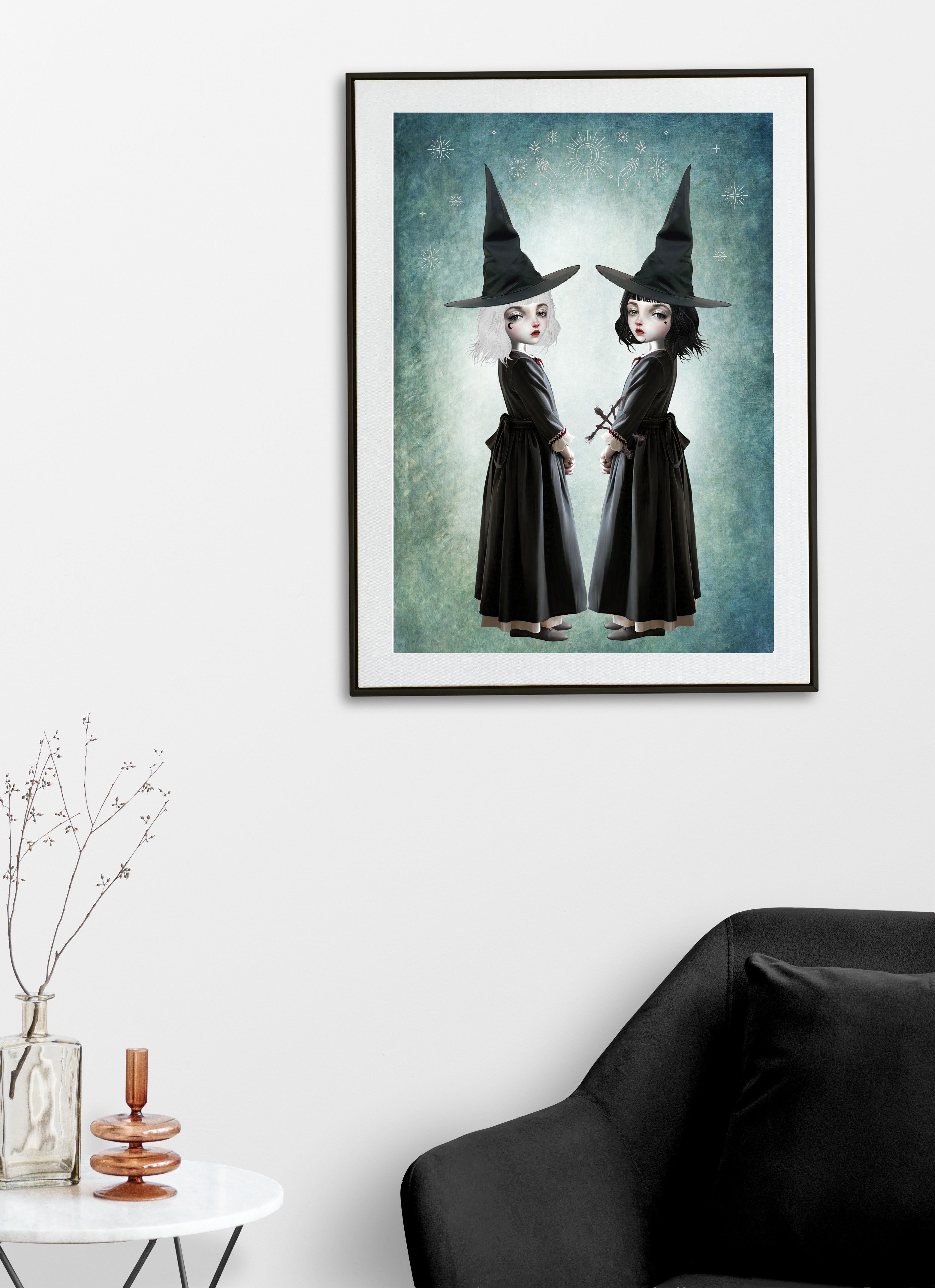 Dark cottagecore fine art print displayed on a living room wall featuring twin gothic witches with white and black hair wearing pointed hats, set against a celestial teal background.