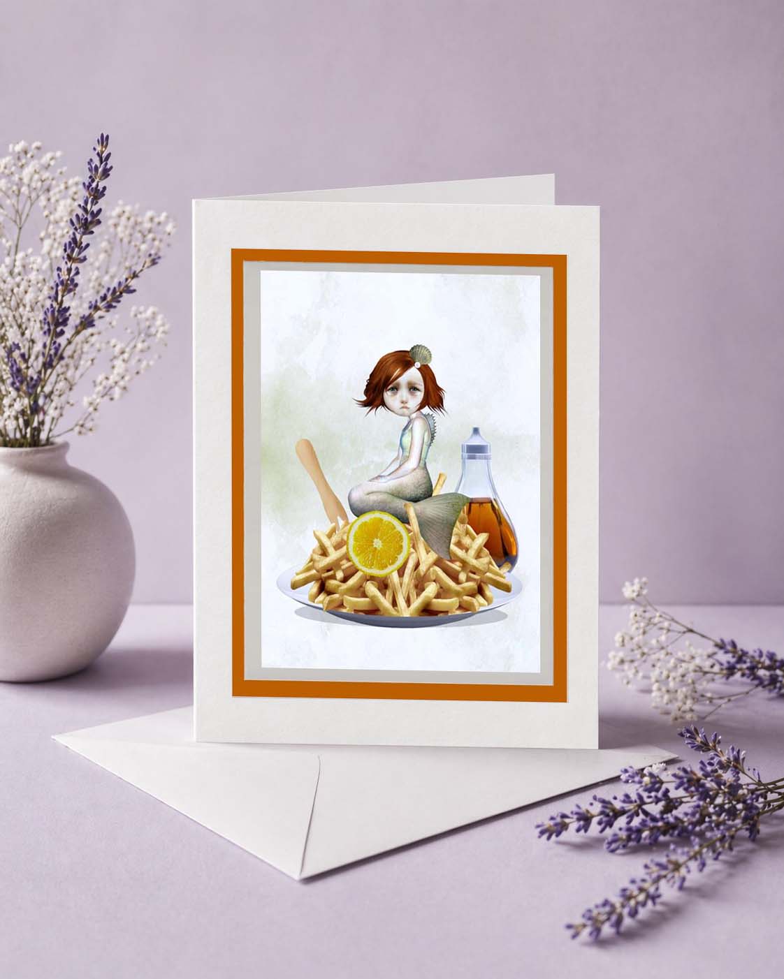 Greeting card with a quirky illustration of a mermaid among fries and a vinegar bottle, placed on a surface with decorative elements.