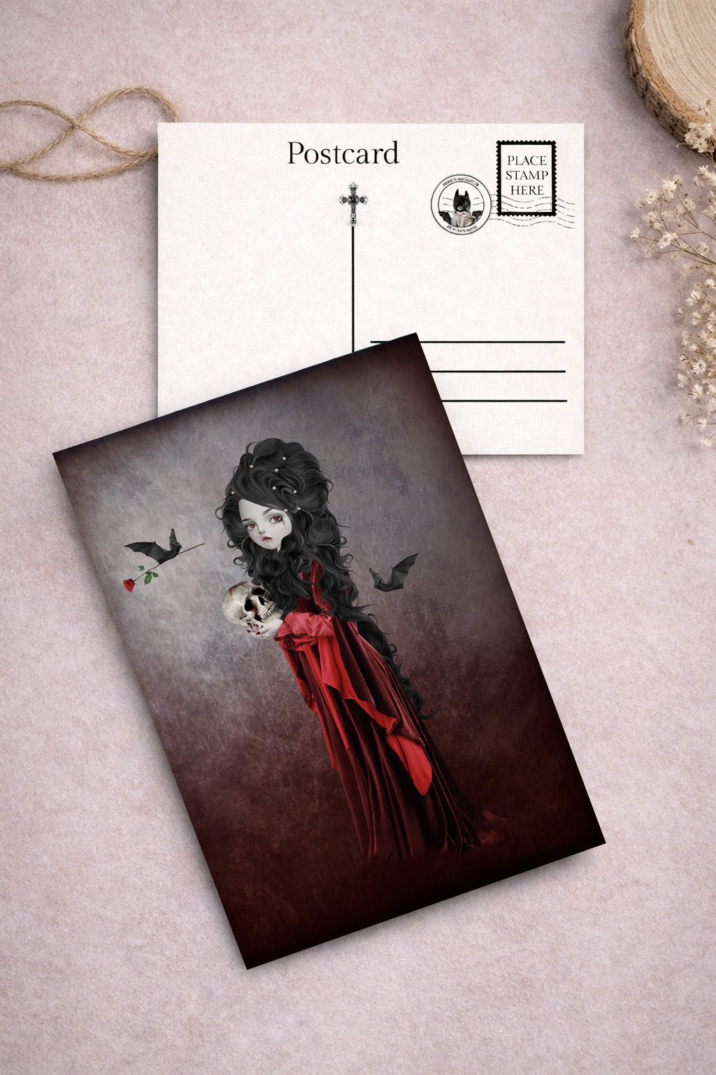 Postcard with macabre design of a vampire in a red dress holding a skull, with a plain postcard on a textured surface.