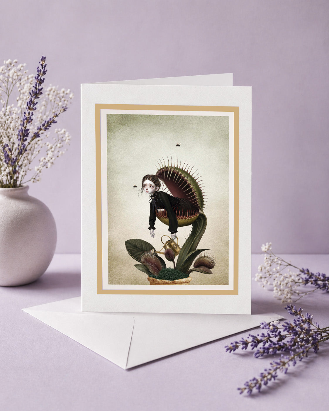 An alternative art greeting card featuring a Victorian gothic illustration of a girl inside a Venus flytrap, displayed upright on its white envelope next to a small ceramic vase with dried lavender and baby's breath flowers on a light purple background.